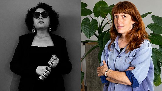 Zwei Frauen im Porträt nebeneinander, links mit lockigem Haar, Sonnenbrille und dunkler Kleidung, rechts mit langen roten Haaren, verschränkten Armen und lockerem Hemd vor großen Blättern.