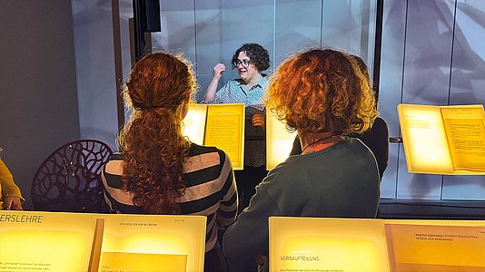Im Vordergrund stehen mehrere Personen mit dem Rücken zur Kamera, die beleuchtete Texttafeln betrachten. Im Hintergrund spricht eine Frau mit lockigem Haar und Brille, die eine gestreifte Bluse trägt. Sie gestikuliert mit einer Hand und steht vor einer hellen Wand mit angedeuteten Schatten. Die Szene vermittelt den Eindruck einer Museumsführung.