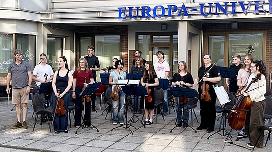 Eine Gruppe von Menschen steht mit Streichinstrumenten und Notenständern im Halbkreis vor einem Gebäude. Die meisten halten Geigen, Bratschen oder Celli, einige tragen lässige Kleidung, andere dunkle Oberteile. Im Hintergrund ist ein Gebäudeeingang mit großen Fenstern und Glastüren zu sehen. Über dem Eingang befindet sich ein blaues Schild mit dem Schriftzug ‚EUROPA-UNIVE'RSITÄT‘. Die Stimmung wirkt konzentriert und freundlich.