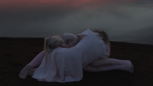 Zwei Personen mit langen, hellen Haaren und weißen, fließenden Gewändern sitzen eng umschlungen am Ufer eines ruhigen Sees. Die Wasseroberfläche spiegelt einen farbenprächtigen Sonnenuntergang mit rosa, orange und violett getönten Wolken wider.