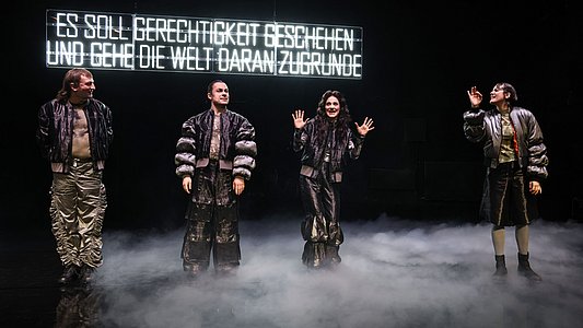 Vier Personen in auffälligen, glänzenden Outfits stehen auf einer Bühne mit Nebel und heben die Hände, im Hintergrund leuchtet der Schriftzug ‚ES SOLL GERECHTIGKEIT GESCHEHEN UND GEHE DIE WELT DARAN ZUGRUNDE‘.
