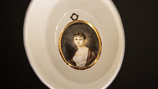 Im Zentrum steht ein rundes Medaillon mit goldfarbenem Rand, das ein Porträt einer Frau mit hellem Kleid und lockigem, hochgestecktem Haar zeigt. Das Medaillon liegt auf einer weißen, leicht gewölbten Unterlage, die von einem dunklen Hintergrund umgeben ist. Oberhalb des Medaillons ist eine kleine Öse sichtbar.