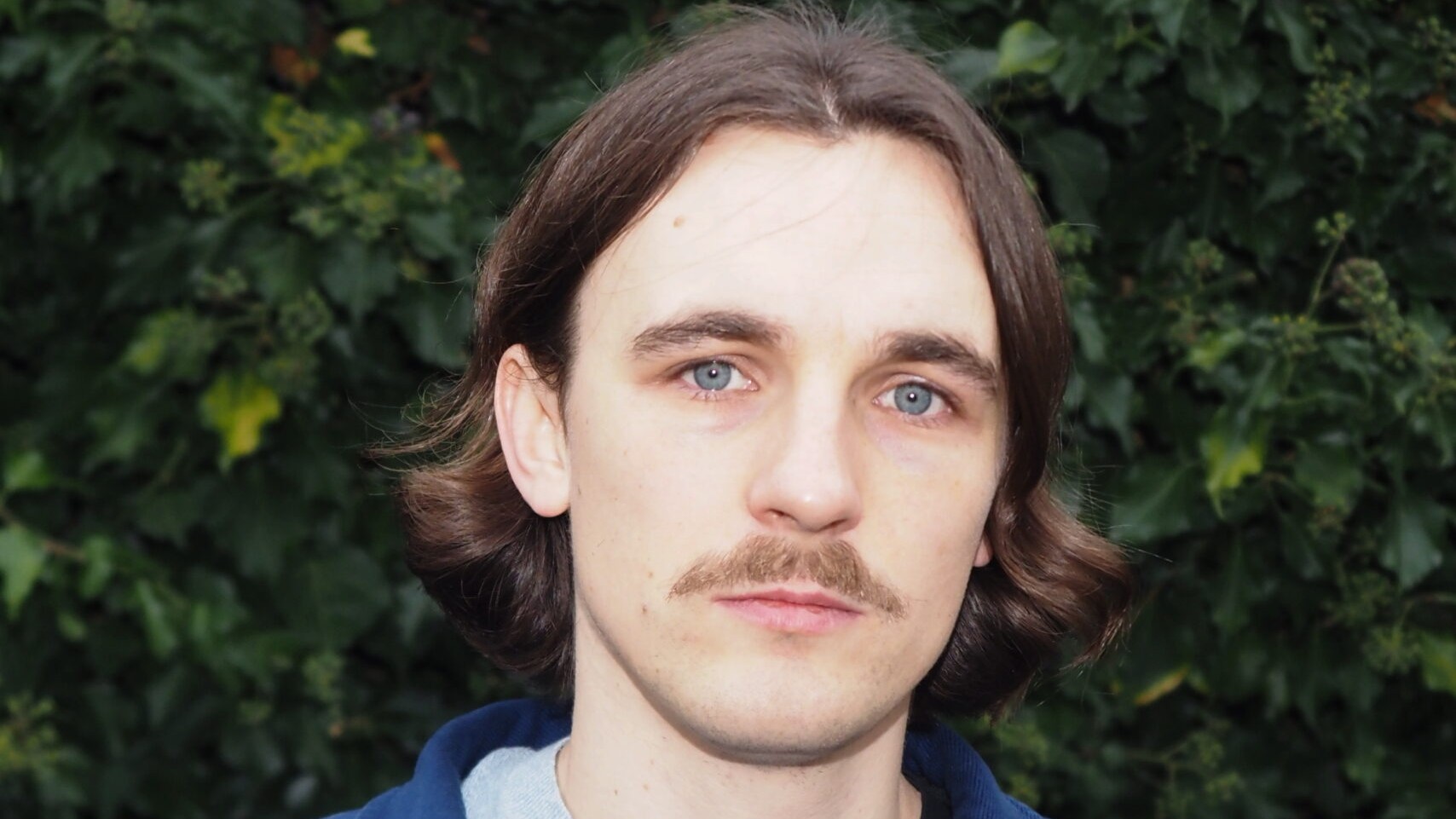 Mann mit mittellangem braunem Haar und Schnurrbart vor einer Hecke