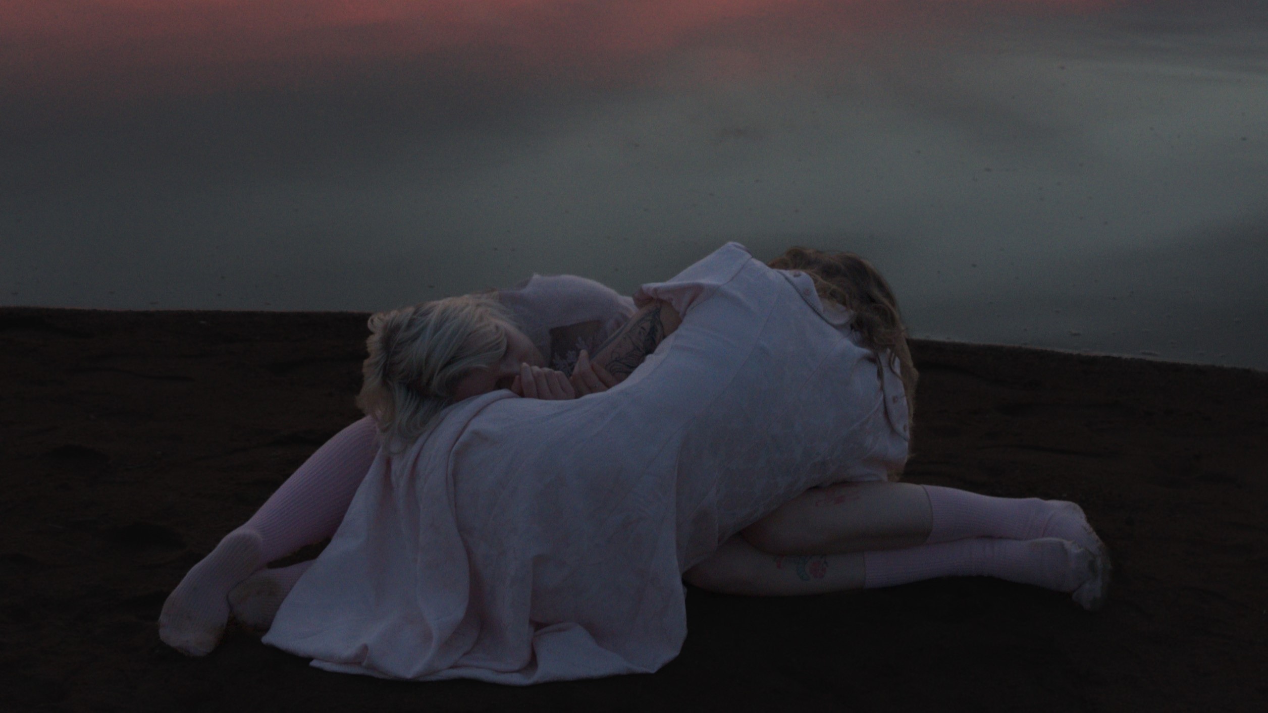 Zwei Personen mit langen, hellen Haaren und weißen, fließenden Gewändern sitzen eng umschlungen am Ufer eines ruhigen Sees. Die Wasseroberfläche spiegelt einen farbenprächtigen Sonnenuntergang mit rosa, orange und violett getönten Wolken wider.